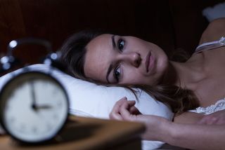 A woman lays awake in bed, looking at a clock.