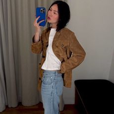 NYC influencer Mimi Nguyen poses for a mirror fitting room selfie in a Madewell store with a bright blue iPhone case, a brown suede jacket, white tee, and jeans