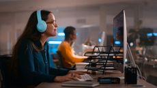Female software developer working on a desktop computer while wearing headphones in an open plan office space with colleague pictured in background.