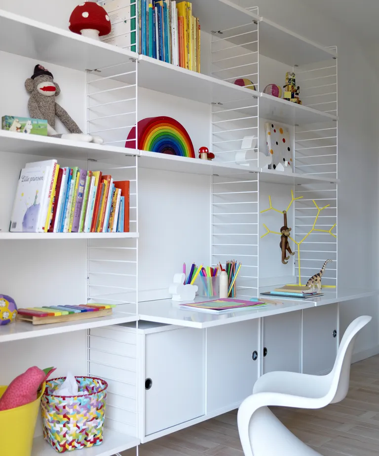 A child's bedroom with white walls, desk and open shelving unit