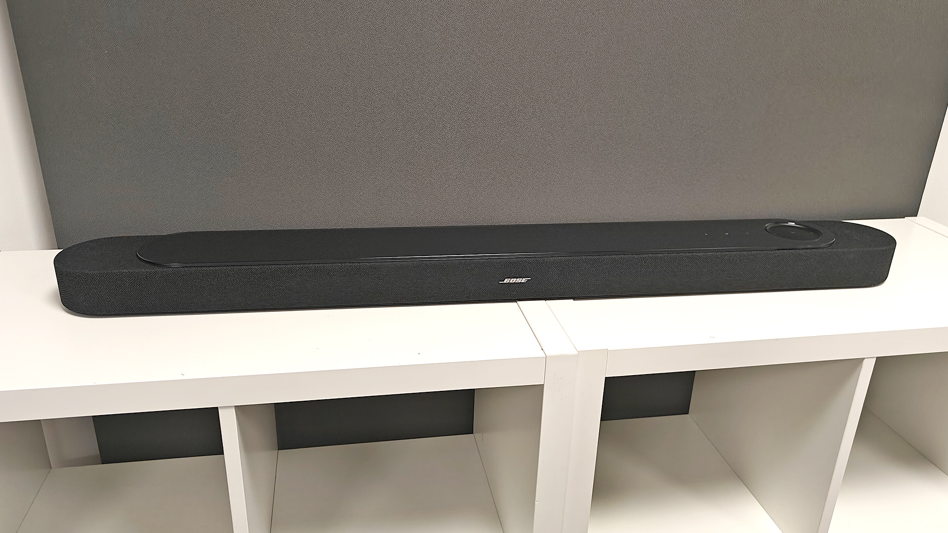 Bose Lifestyle Ultra Soundbar soundbar on white wooden shelving unit in front of grey baffle