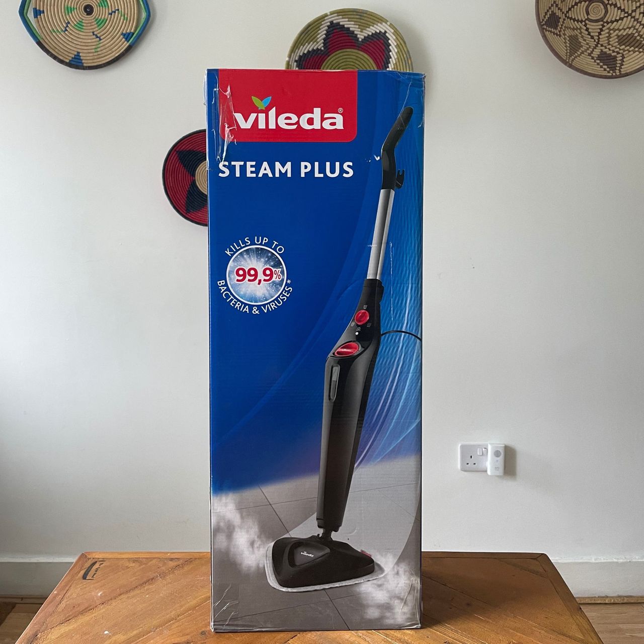 Vileda Steam Mop Plus sitting on a wooden table in front of a white wall with colourful baskets on it