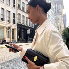 female fashion influencer Tylynn Nguyen poses on the cobblestone streets of the Soho neighborhood in New York City looking at her cellphone wearing a cream jacket, black Savette bag, and Breitling watch