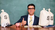 A quirky man sits at a table with bags of money, coins and cash and gives a thumbs-up.