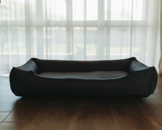 A black leather pet bed on brown wooden flooring with sheer white curtains behind