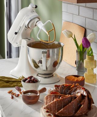 A marble kitchen countertop with a white Kitchenaid, two bowls, a vase of tulips, and a cake on top of it