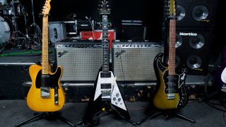 Three electric guitars on a stand in front of some guitar amps