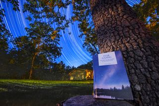 A book leans against a tree against the background of a house in the distance and a dark grassy yard. The night sky is filled with the streaks of stars, traced through the sky through long exposure photos.