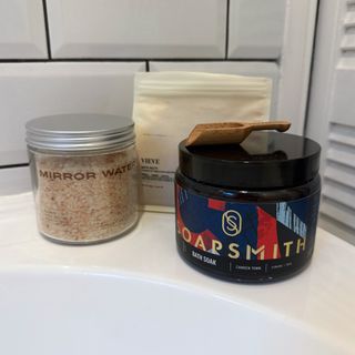 Image showing a close up of three bath salt containers from (left to right) Mirror Water, VIEVE and Soapsmith, balanced on the corner of a white bathtub, with white tiles and a cupboard door in the background