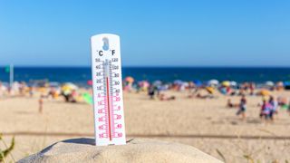 A thermometer standing in a mound of sand at a crowded beach