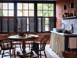 Dark colored window shutters are seen in a rustic kitchen with open shelving. A dining table sits in the middle of a room with an open book, candle and other items