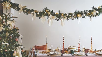 A dining room with a set dining table for Christmas with a Christmas tree to the side and a garland above decorated with white bows