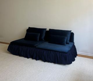 Image of a velvet, navy sofa with a ruffle skirt in a room that has white walls and a large white area rug