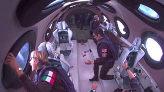 four people wearing dark flight suits float inside a space plane's white-walled cabin.