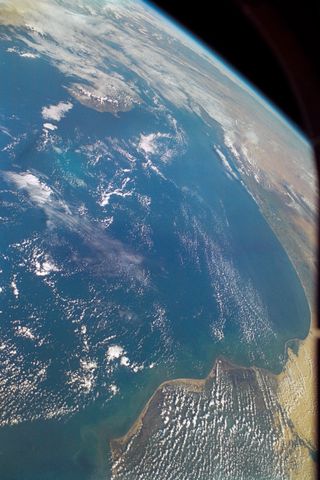 Vast swathes of blue ocean and thin wispy white clouds along with yellow land to the right.