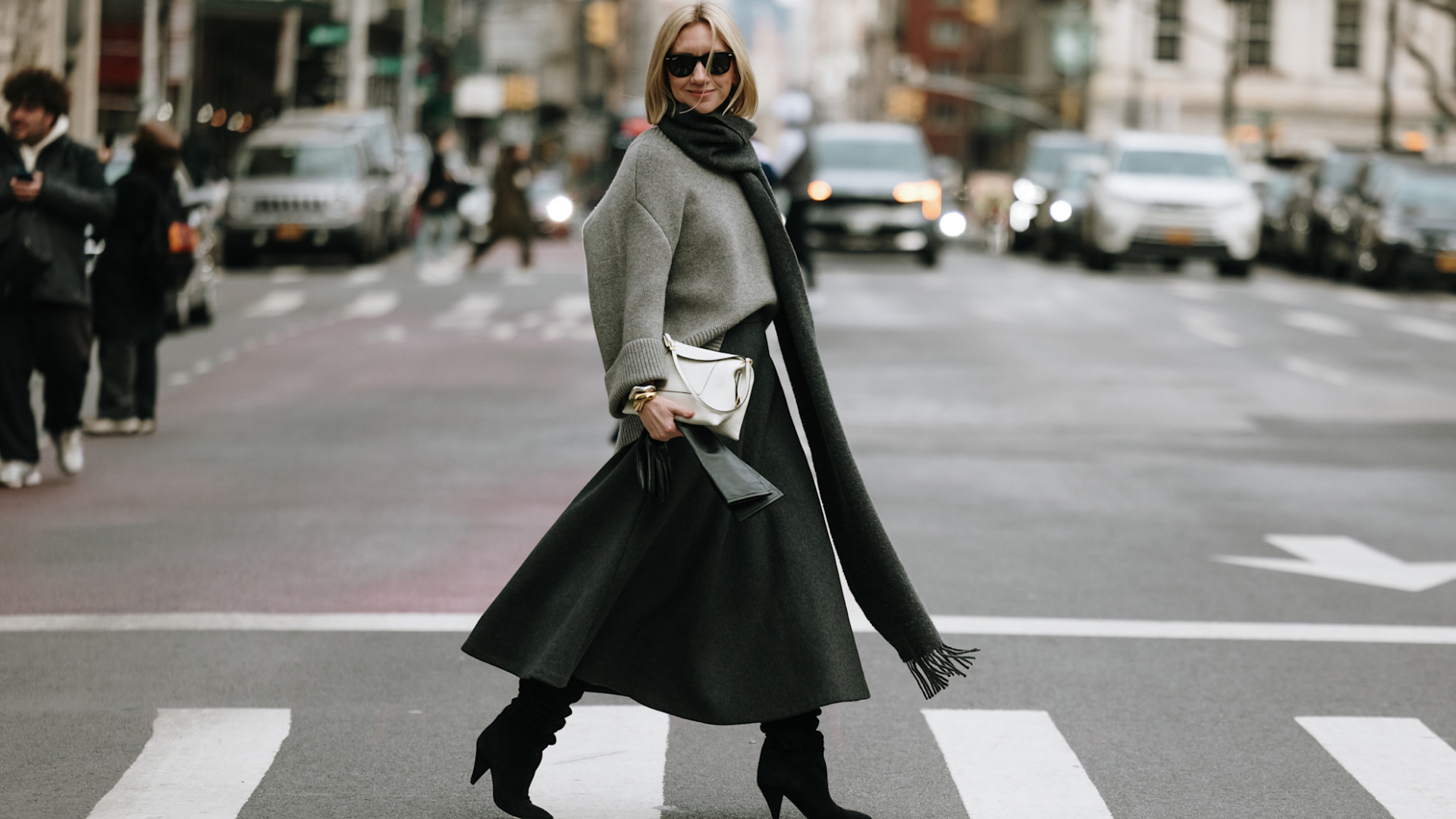 Woman crossing the street wearing sunglasses big scarf, grey sweater, white bag, and black skirt 