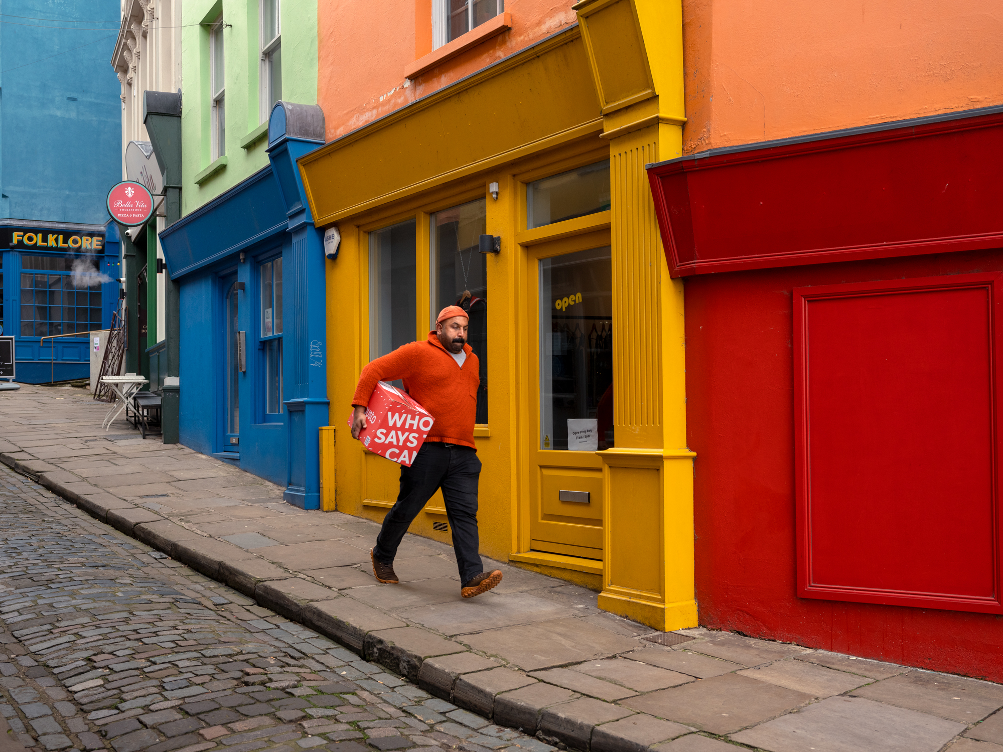 A man wearing a bright orange sweater and cap walks down a cobblestone sidewalk carrying a red box past a row of vibrant, colorful storefronts.