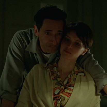 adrien brody leans over felicity jones as they look over blue prints in a still from the brutalist