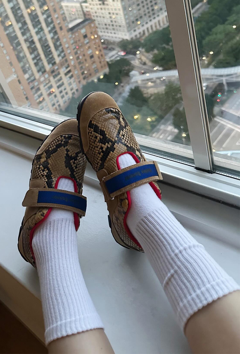 The best ballet sneakers are shown in a close photo of a woman's feet on top of the ledge of a window wearing long white socks and Mary Jane ballet sneakers with snakeskin print