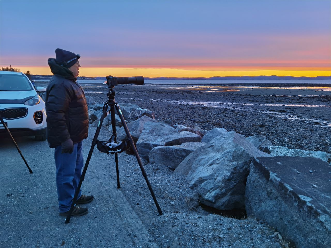 a man stands next to a camera and a tripod looking out to a cloud laden sky.
