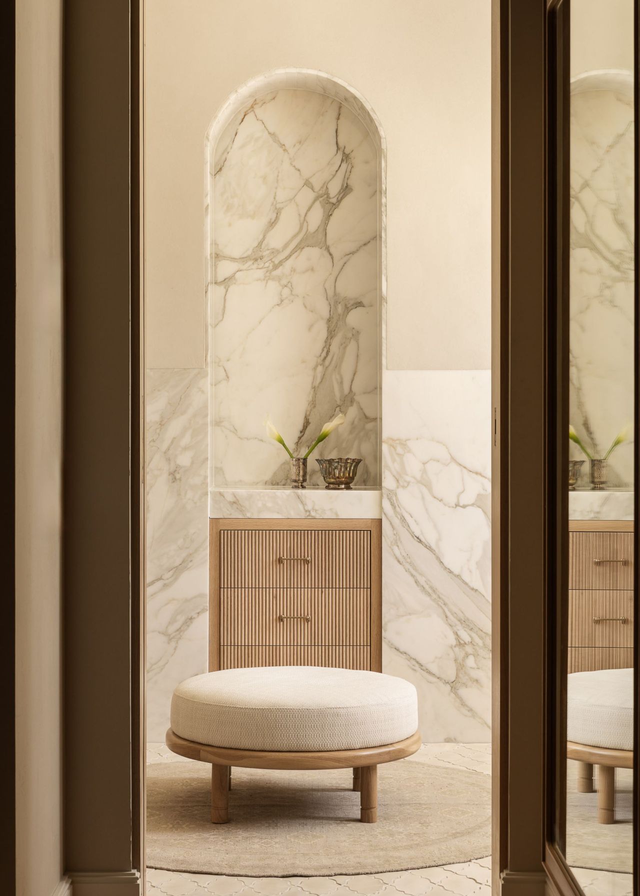 A modern neutral bathroom with cream walls and marble, an arched alcove with storage and two minimal decor pieces on top in front of a round cream-cushioned, wood-based footstool on a round cream rug