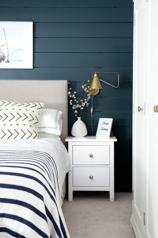 Bedroom with blue panelled walls, beige upholstered bed, blue and white striped bedding and brass wall light