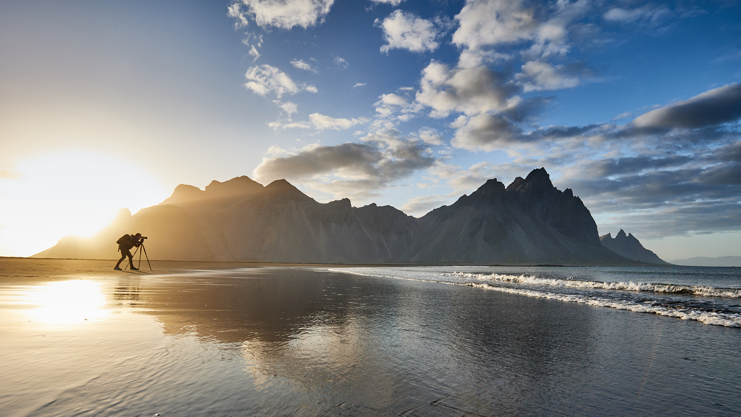A photographer captures the sunset on a beach with dramatic mountain silhouettes reflecting in calm water