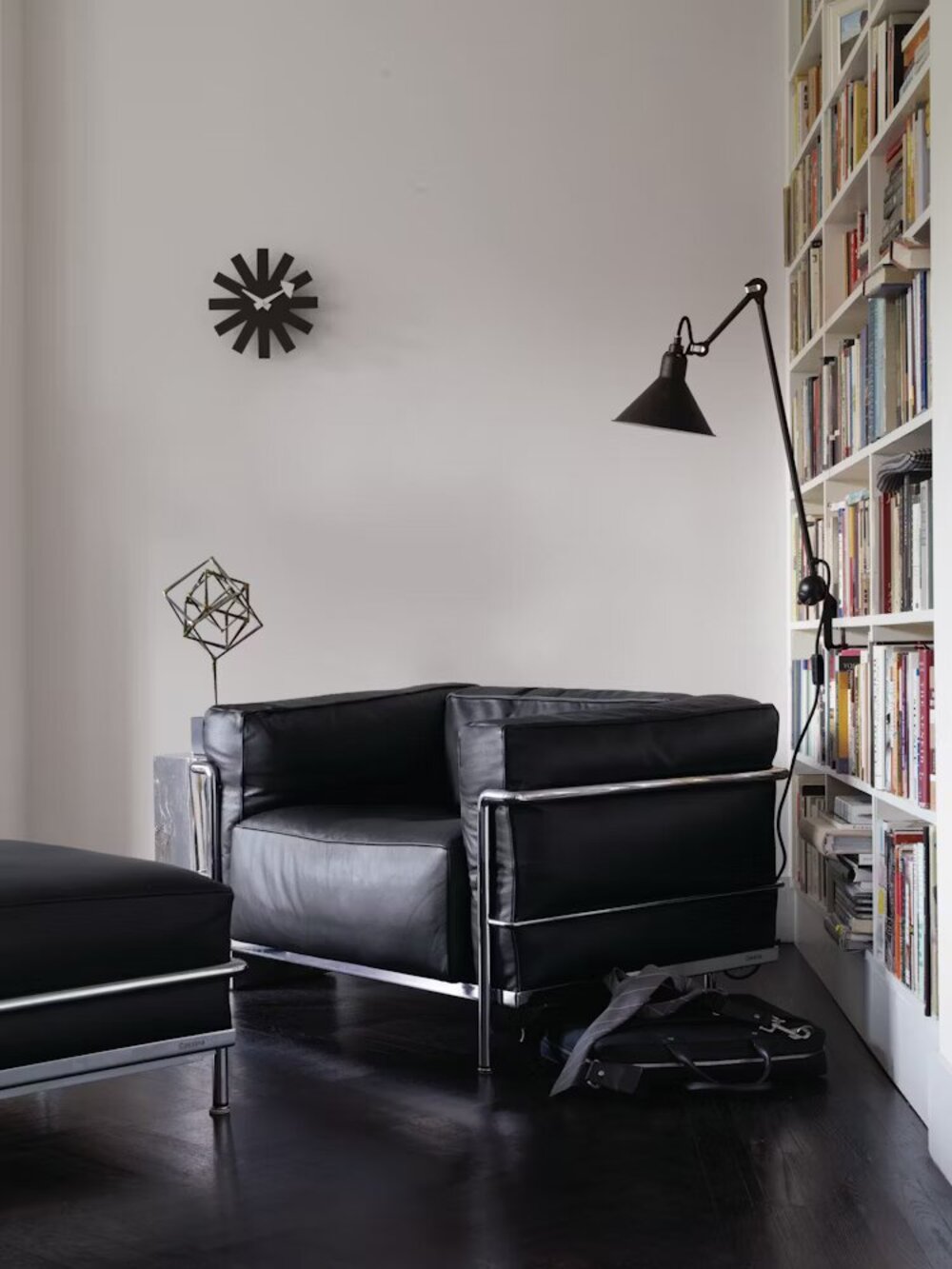 living room with leather armchair, bookshelf, lamp attached to bookshelf, leather ottoman, dark floorboards, and wall clock