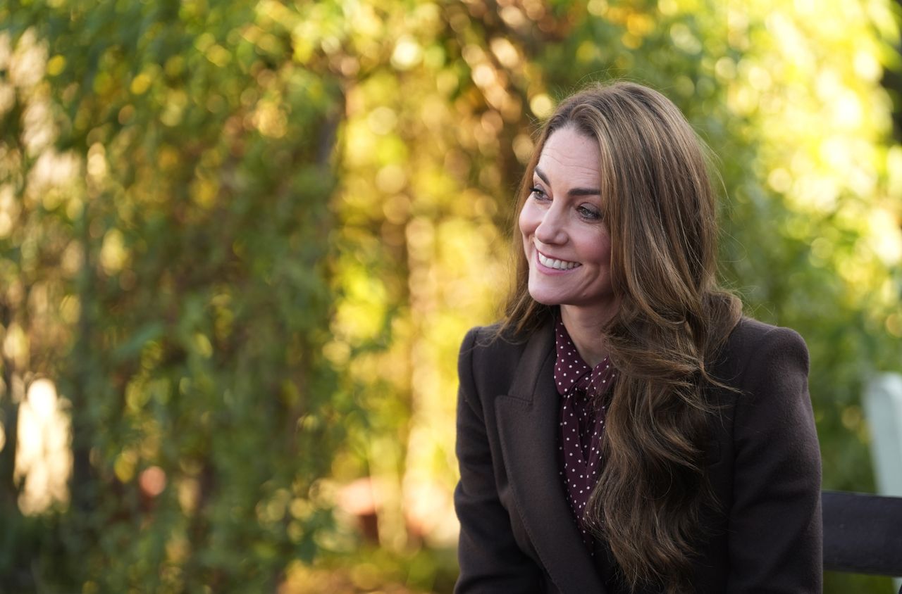 Kate Middleton wearing a brown coat and burgundy polka dot dress smiling with green trees behind her