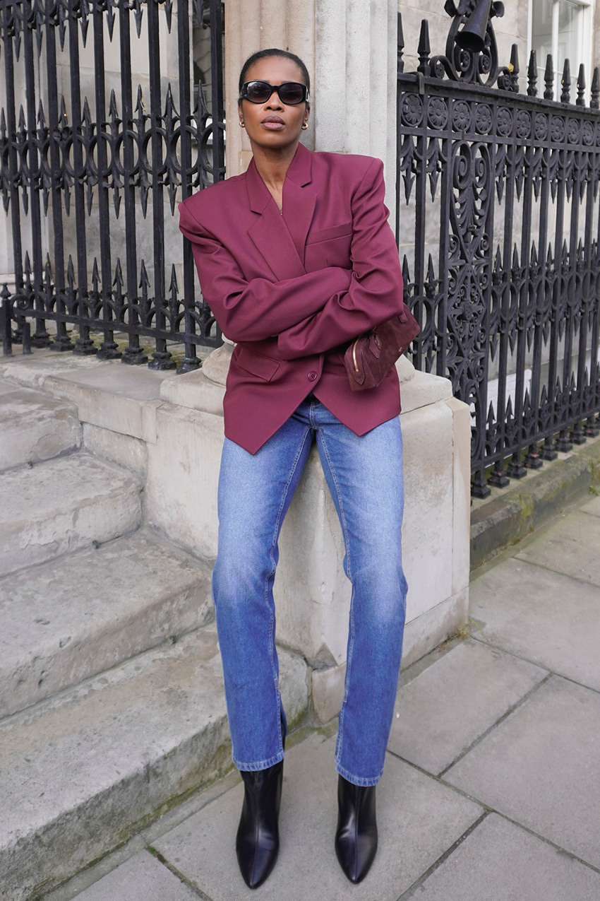 How to wear skinny jeans is shown in a photo of a woman leaning against a column on the sidewalk wearing black sunglasses, gold hoops, a red blazer, a red belt, a red suede shoulder bag, skinny jeans with a split hem, and black ankle boots