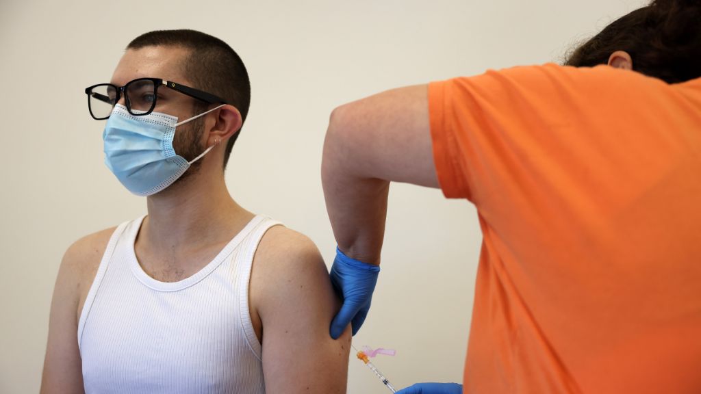 a nurse wearing a orange shirt administers a vaccine to a young man wearing a surgical mask and white tank top