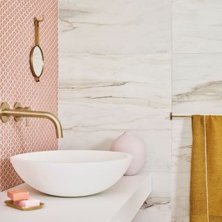a bathroom with white marble tile wall and a pink mozaic tile wall backsplash behind an above counter white bowl sink with gold chrome tap fixings