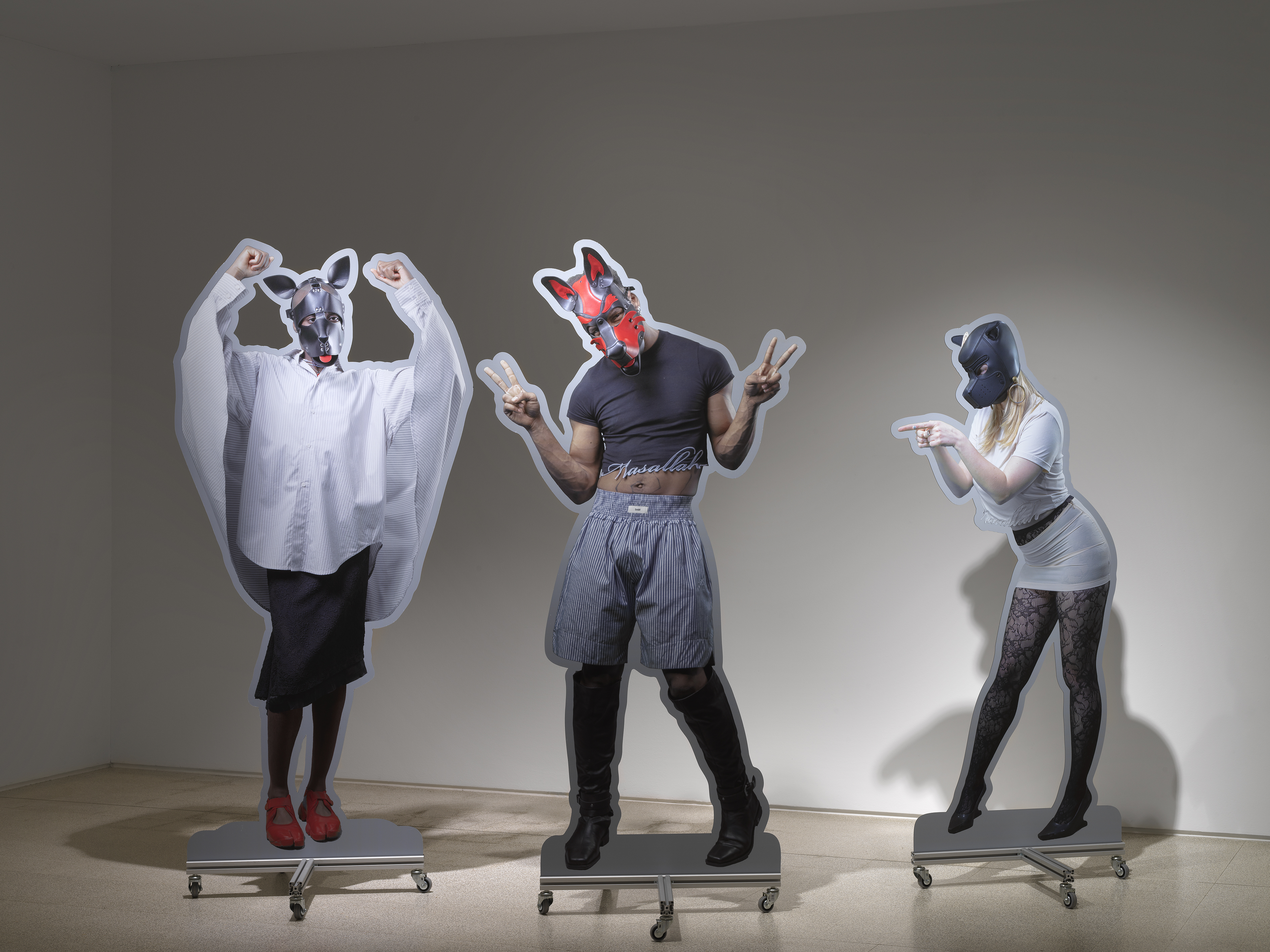 Three life-size photographic cutouts of dog-masked figures mounted on wheeled steel plinths in a gallery space: from left, a figure in an oversized white shirt with arms raised; centre, a muscular figure in shorts and knee-high boots making peace signs; right, a figure in a white dress and patterned tights pointing outward.