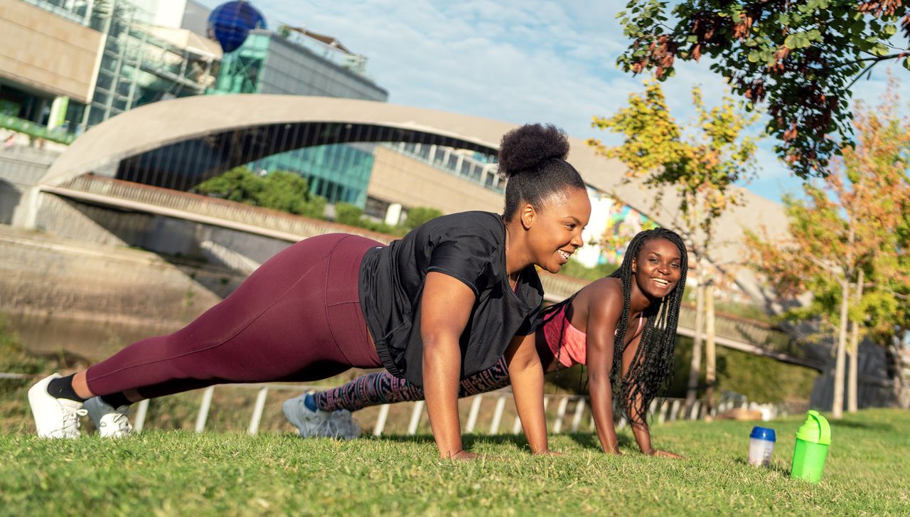 Two women outside in a park in the push-up position, supported on their hands and toes. They are both smiling and wearing sportswear