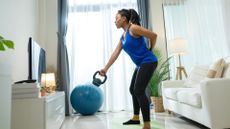 woman wearing blue vest and dark leggings in her living room. facing sideways to the camera holding a kettlebell with one hand doing a swing. 