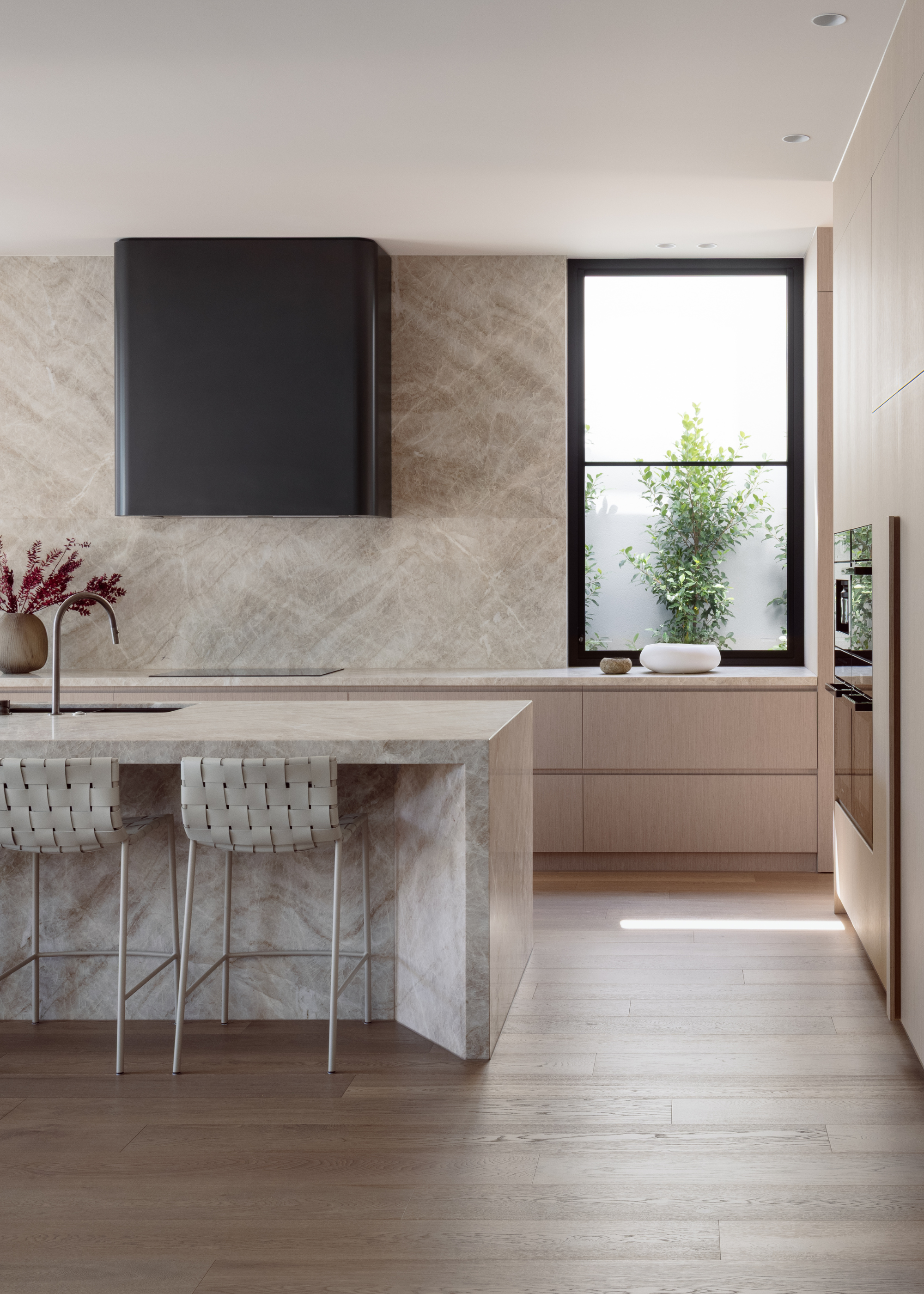 A bright minimalist kitchen with a dark gray range hood, a stone counter and backsplash, a stone island, and a pair of woven leather stools
