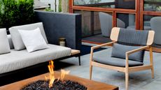small outdoor area with gray pavers, rusty firepit, timber armchair with gray upholstery, and a slim-line, low-profile outdoor sofa with light gray upholstery and a planter box behind it