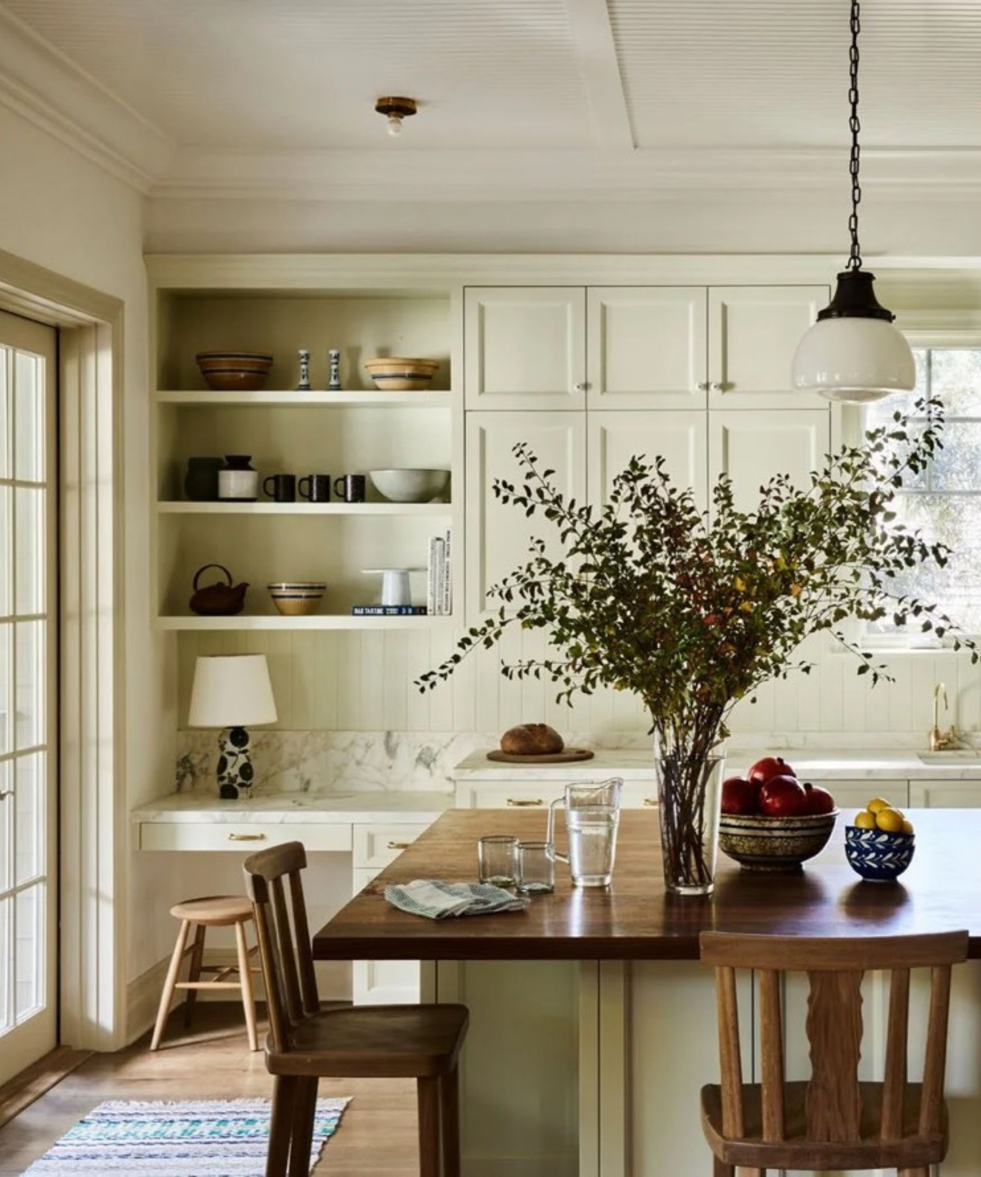 A bright, airy kitchen with cream-colored cabinetry, a large dark wood island with barstool seating, and marble countertops. Two white pendant lights hang above the island, and a large vase of greenery sits as a centerpiece near a window.