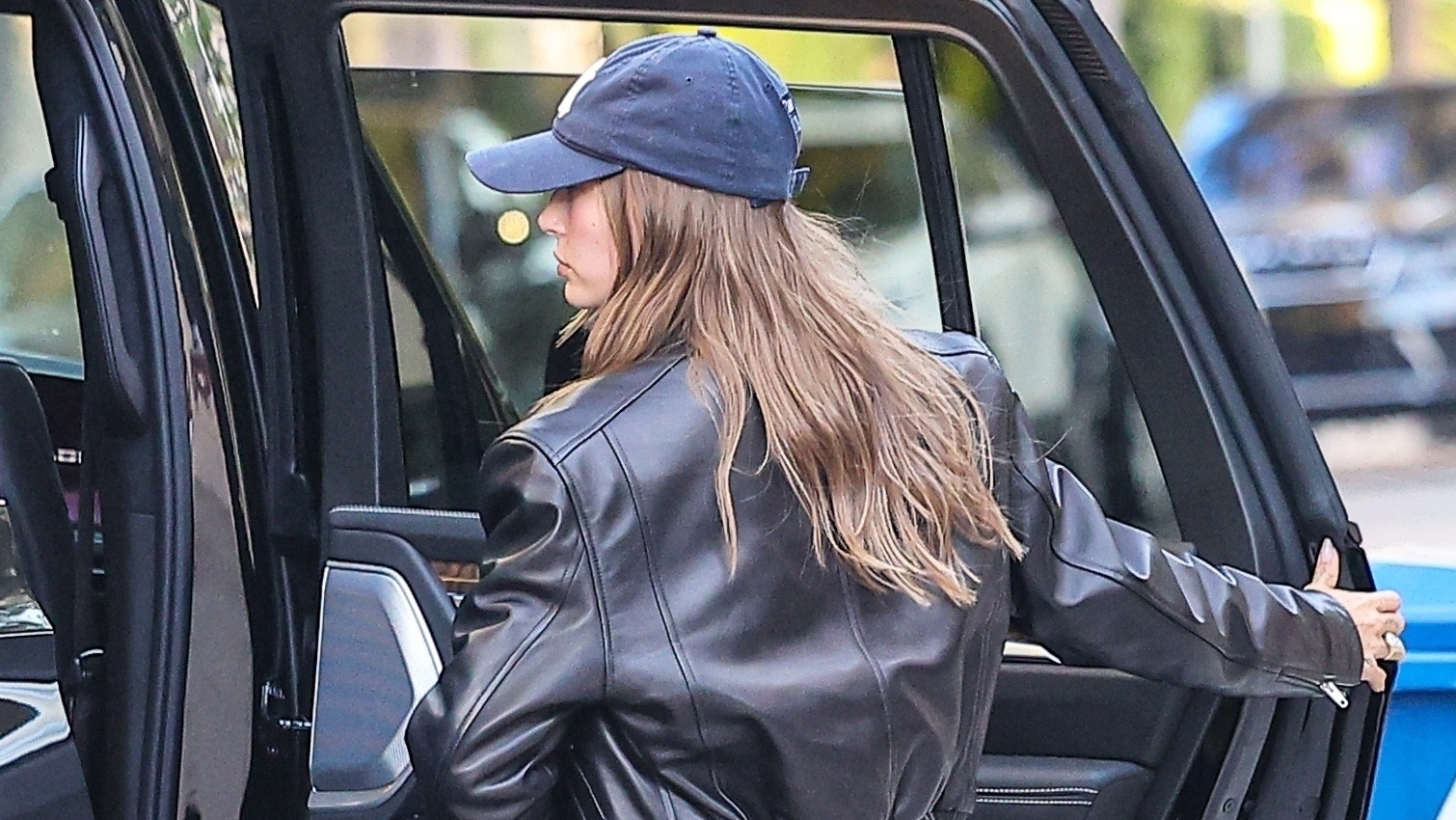 Hailey Bieber wearing a NY Yankees baseball cap, leather jacket, black pull-on pants, and flip-flops 