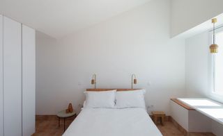 Interior view of a room at Casa Modesta featuring white walls, floor tiles and a bed with a wooden headboard, two lamps, white pillows and white linen. There are also two different bedside tables, a white wardrobe and a pendant light by the window