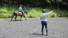 A riding instructor pictured from behind gesturing with her right hand as a adult rider on a bay horse canters on the right rein.