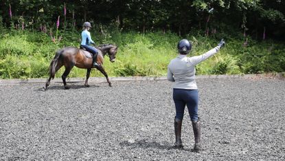 A riding instructor pictured from behind gesturing with her right hand as a adult rider on a bay horse canters on the right rein.