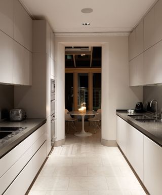 A modern galley kitchen with sleek, handleless white cabinetry on both sides. Black countertops contrast with the white units. A built-in hob sits flush on the left-hand counter, with a toaster nearby. A black faucet and sink are on the right-hand counter.