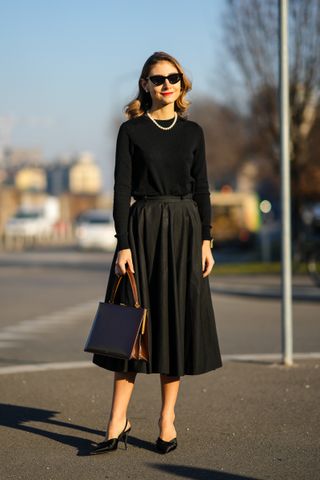 Woman posing and wearing a black top and full midi skirt, black slingback heels, sunglasses, and a structured bag