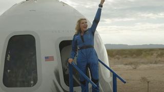 a woman in a blue flight suit pumps her hand in the air while standing in the doorway of a white cone-shaped capsule