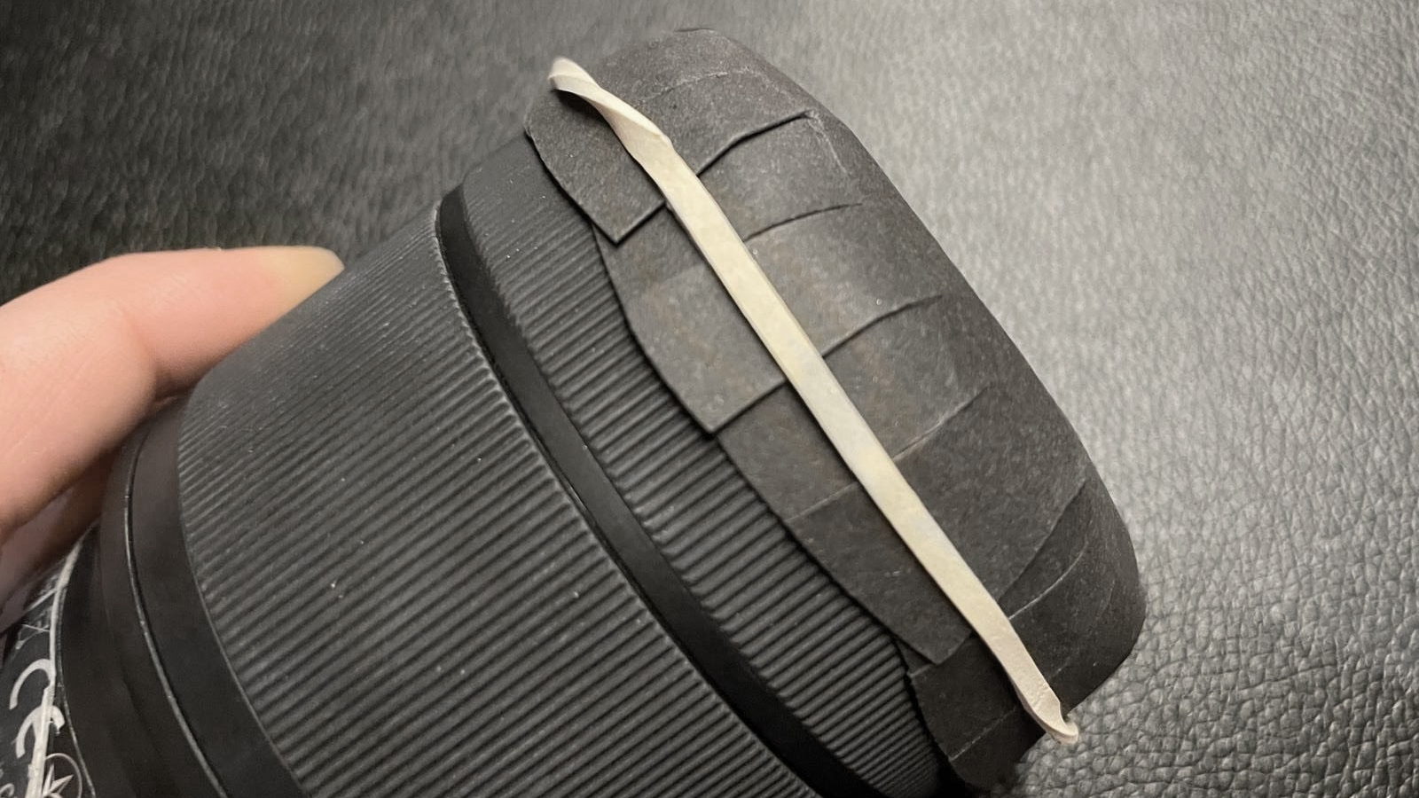A person's hand holds a black cylindrical object wrapped with a rubber band, showing textured grip and a patterned base on a dark surface