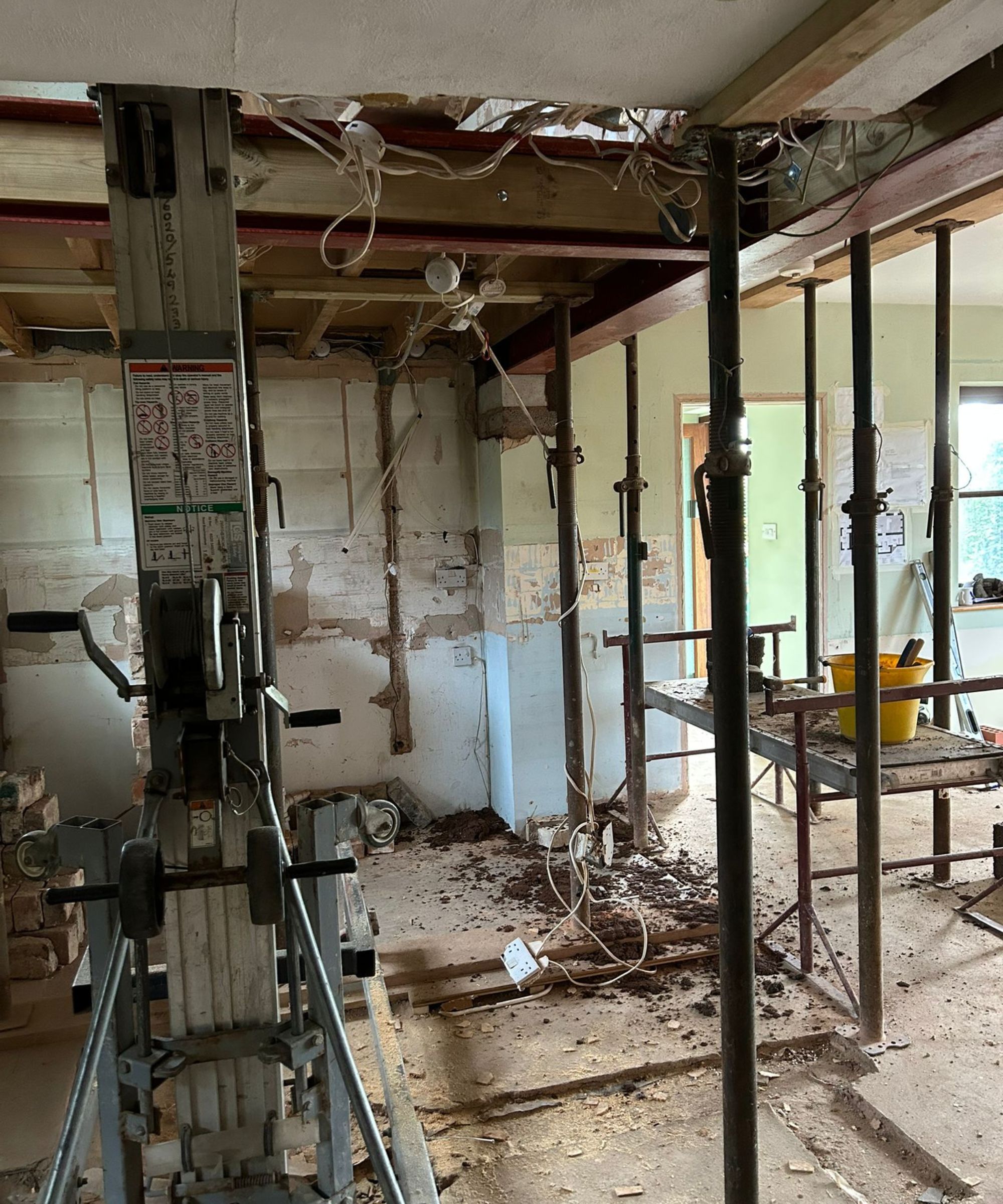 A heavily stripped room supported by multiple acrow props and steel beams. Exposed wiring hangs from the ceiling, while a mechanical lift and scattered debris fill the space. The walls show layers of removed plaster, revealing the structure beneath.
