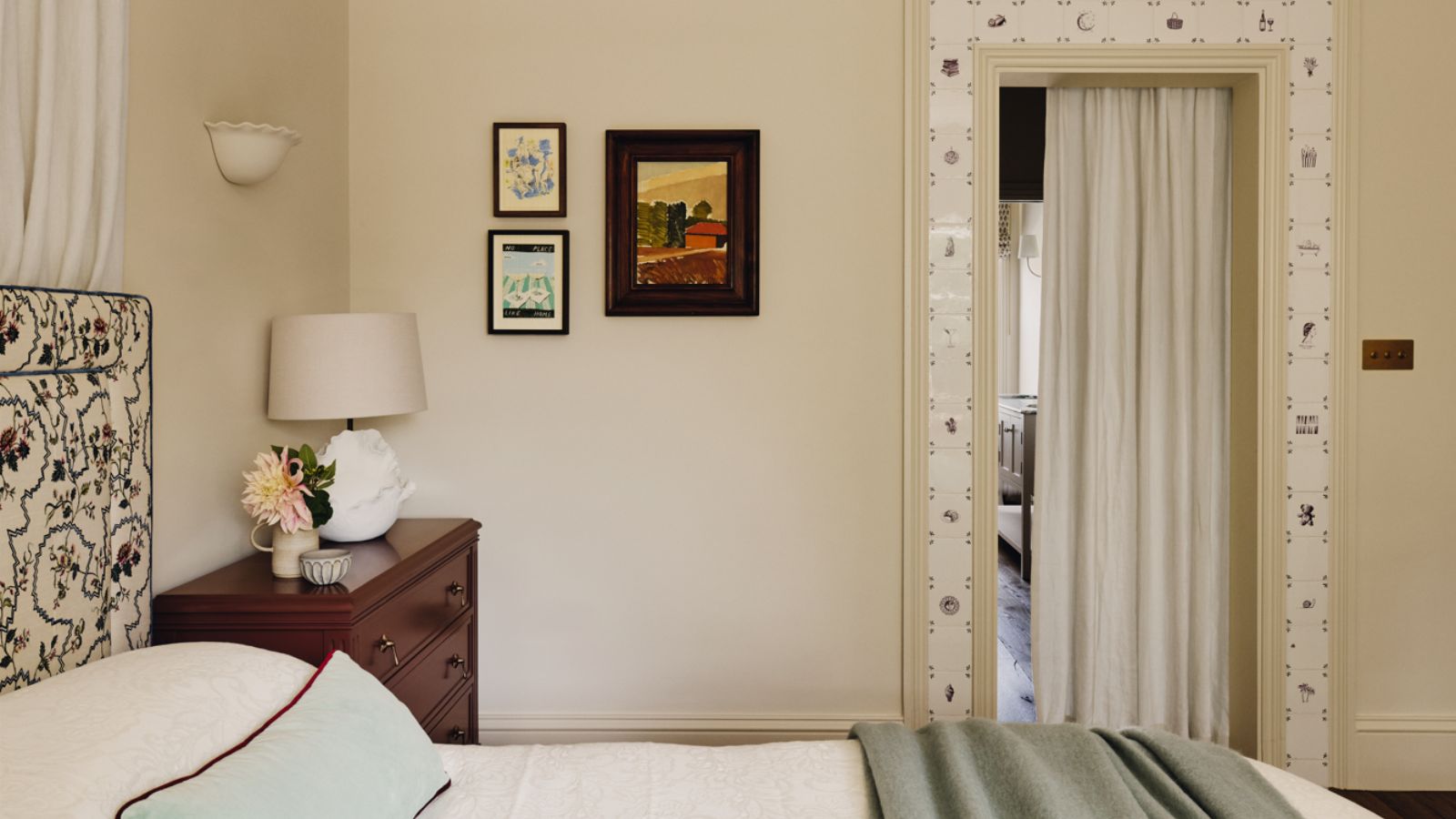 neutral bedroom painted in Farrow &amp; Ball's Dimity with delft tile door trim, small vintage artwork, a floral upholstered headboard and a burgundy nightstand