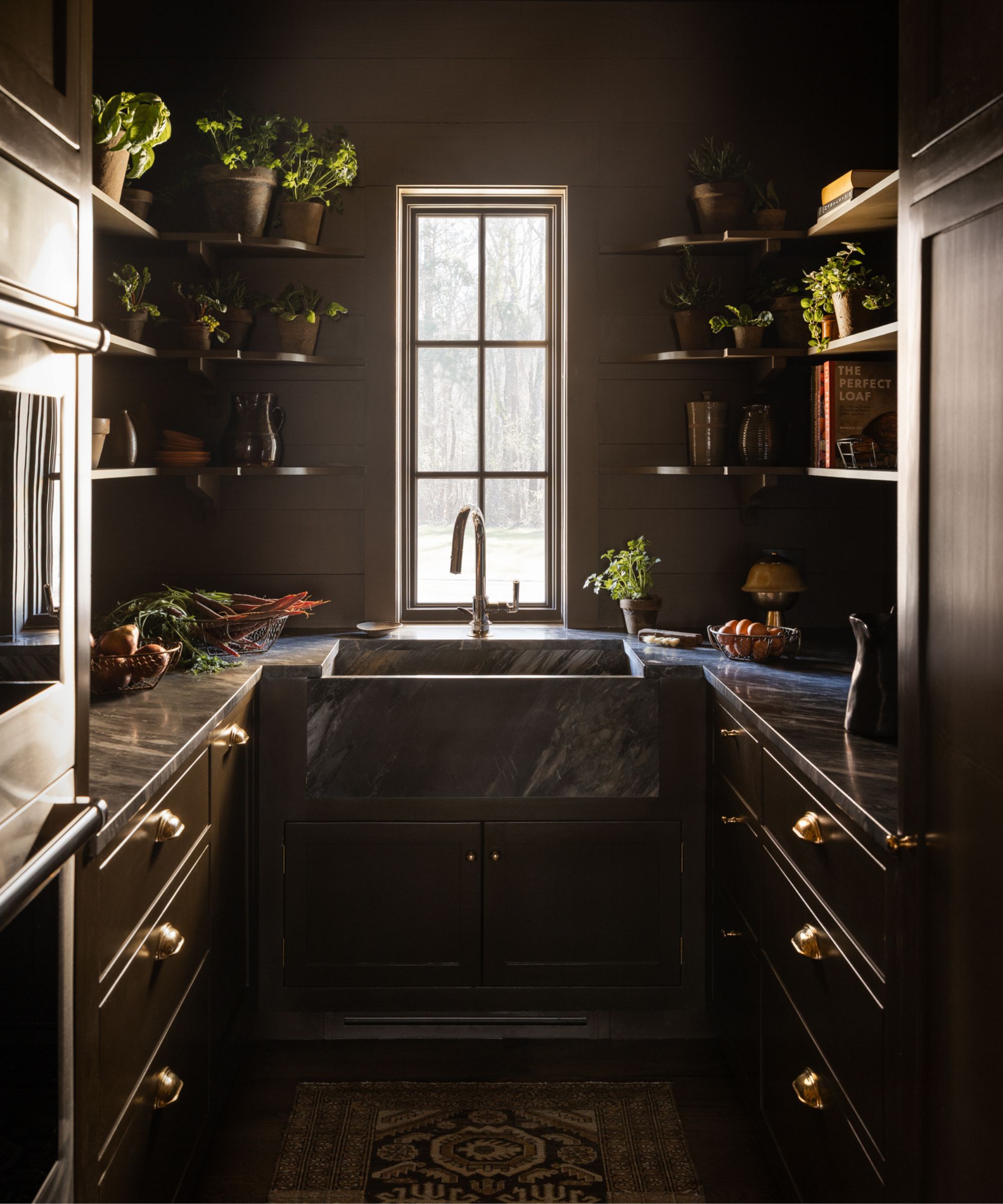 Hidden working kitchen with dark cabinetry, marble countertops, open shelves, and a farmhouse sink beneath a window.