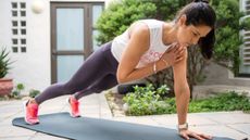 A woman is performing a plank tap outside on a yoga mat. She is balancing on her left hand, with her left arm straight on the ground, and both legs extended behind her so that her elevated body forms a straight line from her shoulders to her heels. Her right hand taps her left shoulder.